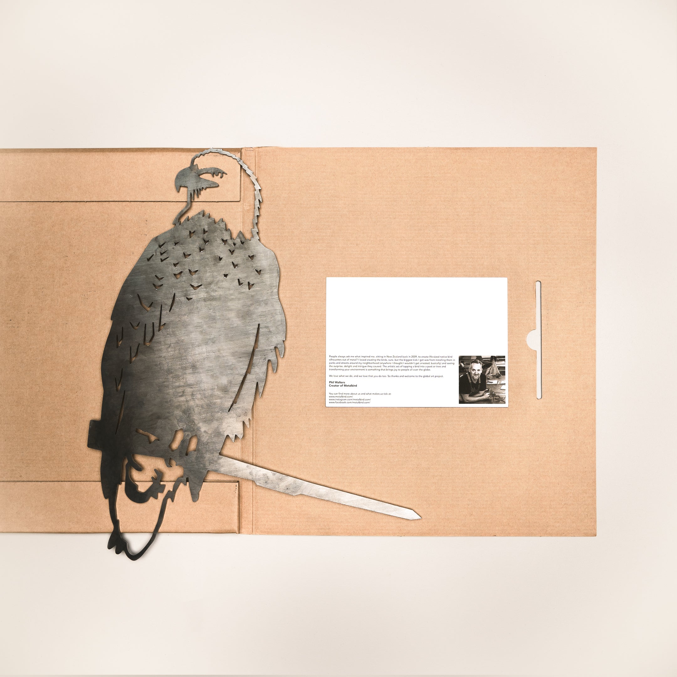 A Metal Bird Garden Art piece featuring a cardinal, hummingbird, or owl rests on a brown cardboard folder beside a white card—perfect as a unique gift for bird lovers or as striking garden décor.