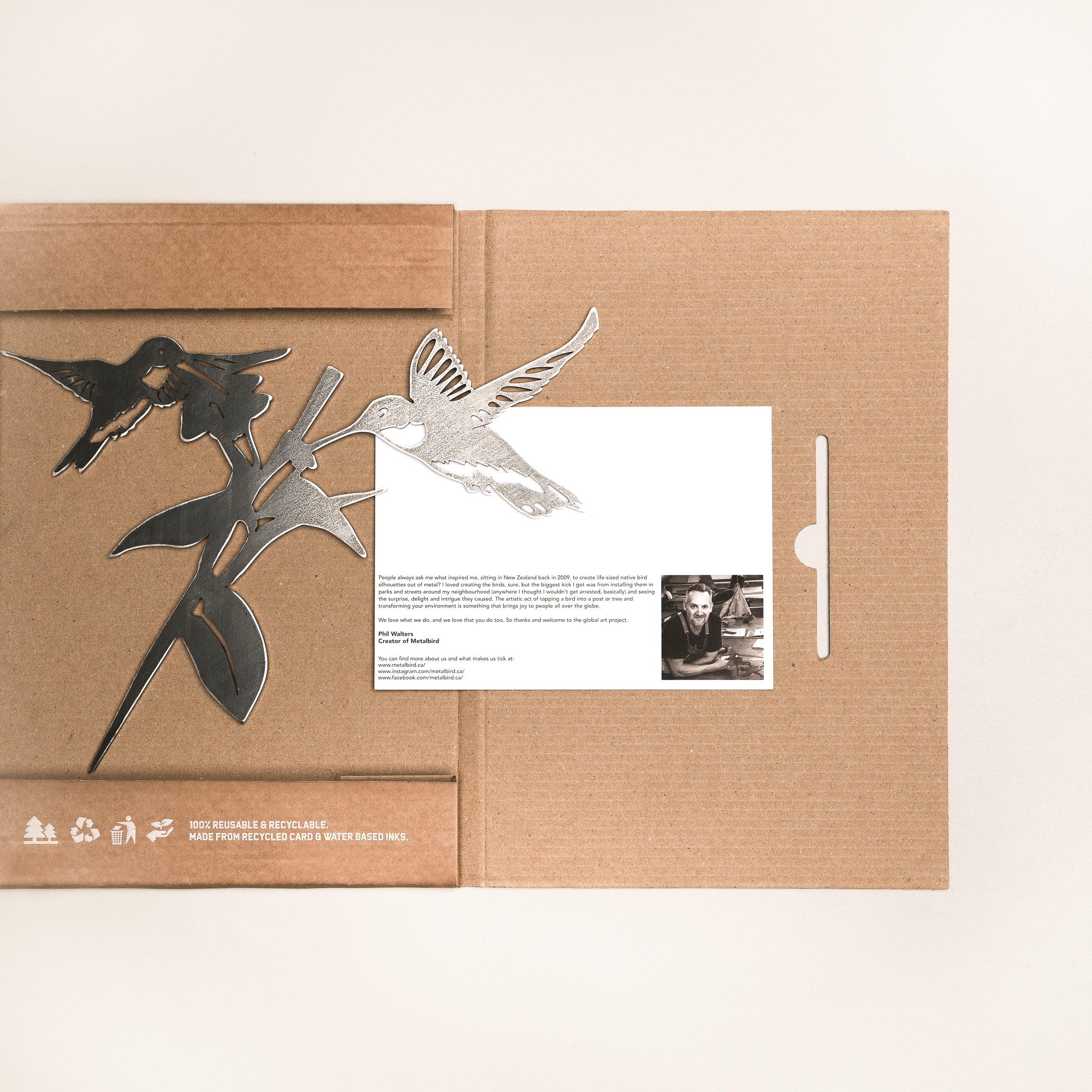 An open cardboard package displays a Metal Bird Garden Art hummingbird sculpture. Included is a white card with text and a black-and-white photo. The scene features a light, minimal background—perfect for bird lovers.