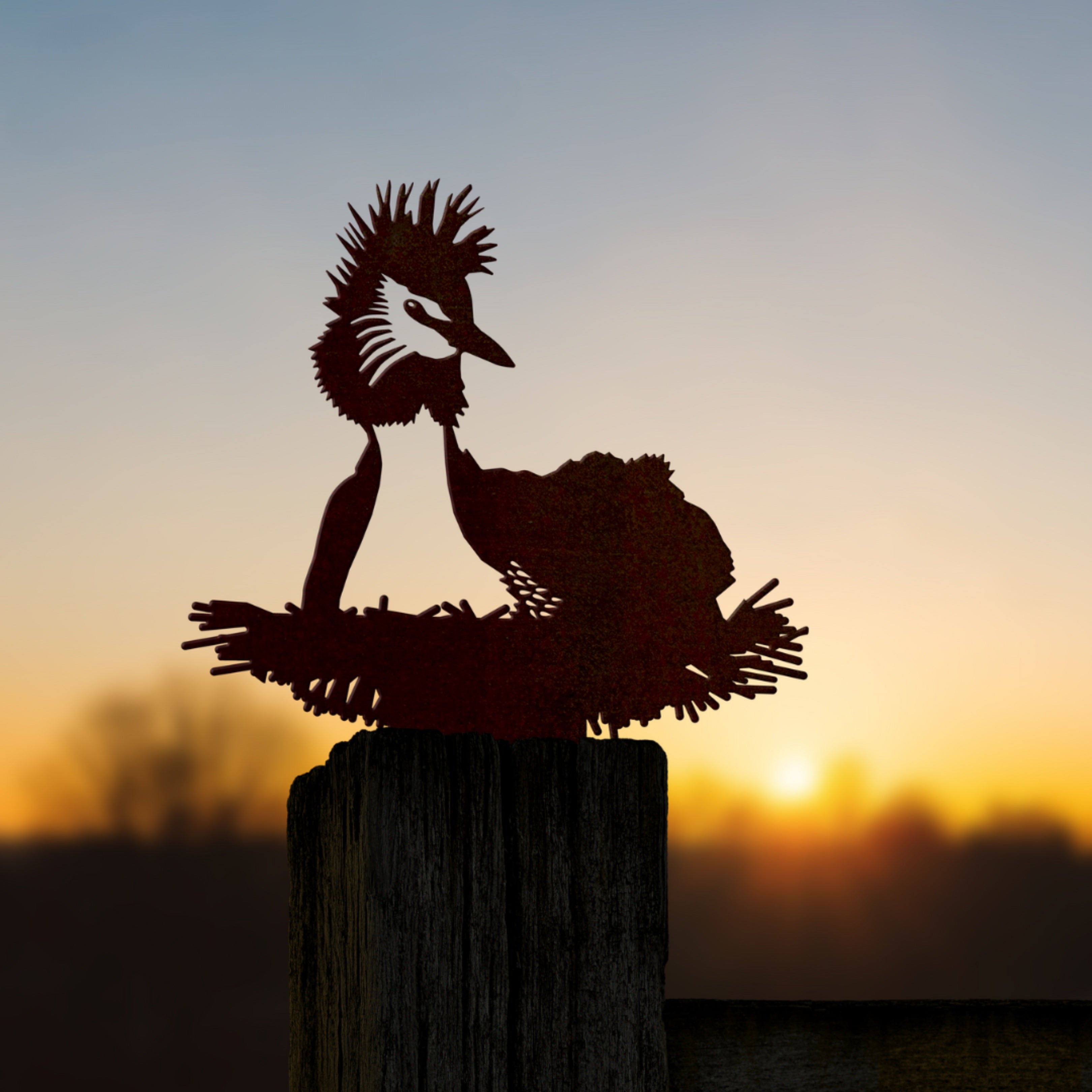 A corten steel Pūteketeke / Crested Grebe decor sits on a fence post, silhouetted against a blurred sunset and trees—an elegant yard art piece.