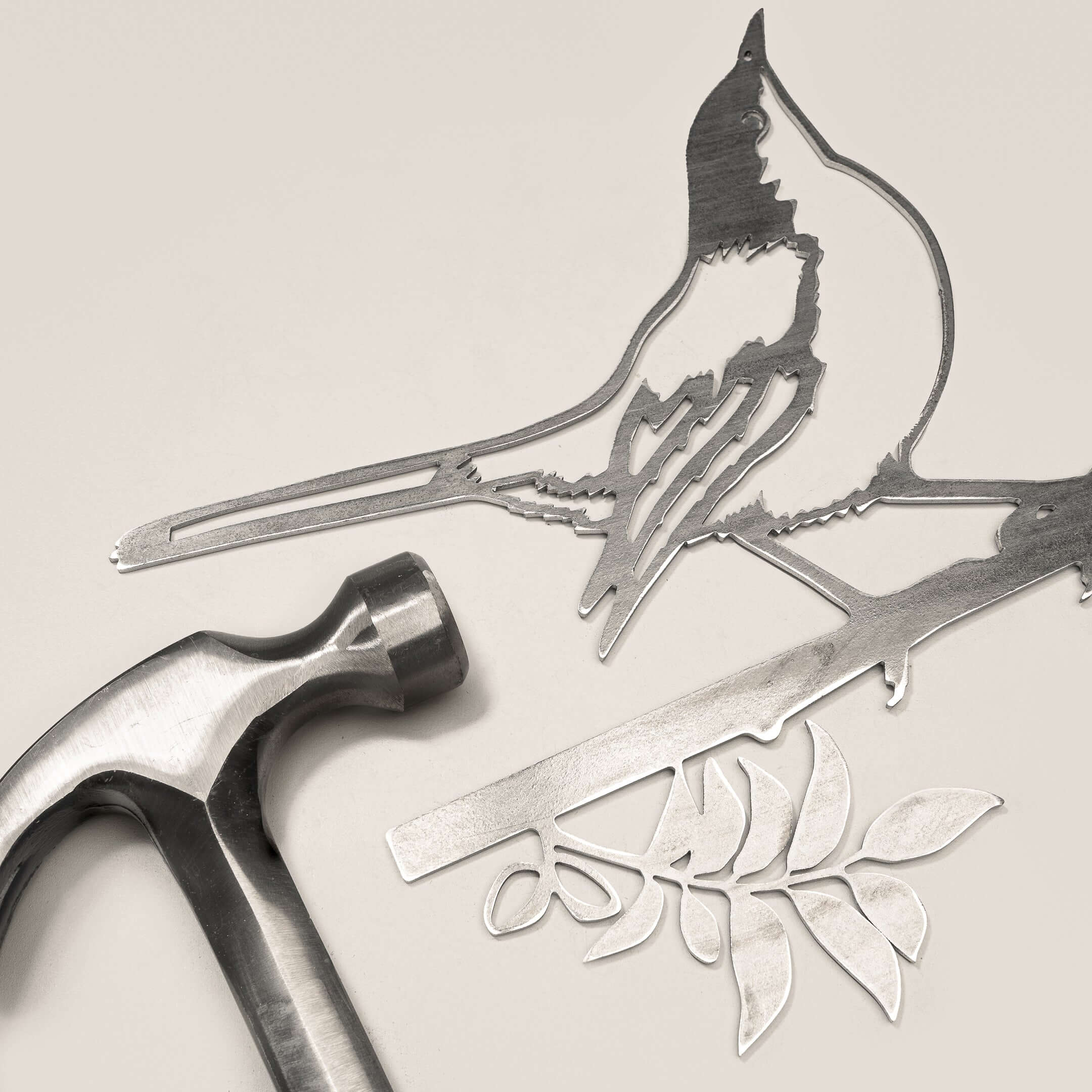 The Mockingbird, a flat metal artwork of a bird perched on a leafy branch, is displayed next to a premium hammer—ideal as elegant yard art or a thoughtful gift, all set atop a light-colored surface.