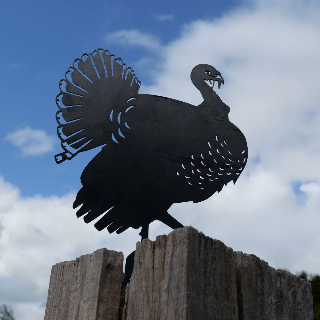 The Turkey features a premium metal silhouette mounted on a weathered wooden post, set against a partly cloudy blue sky.