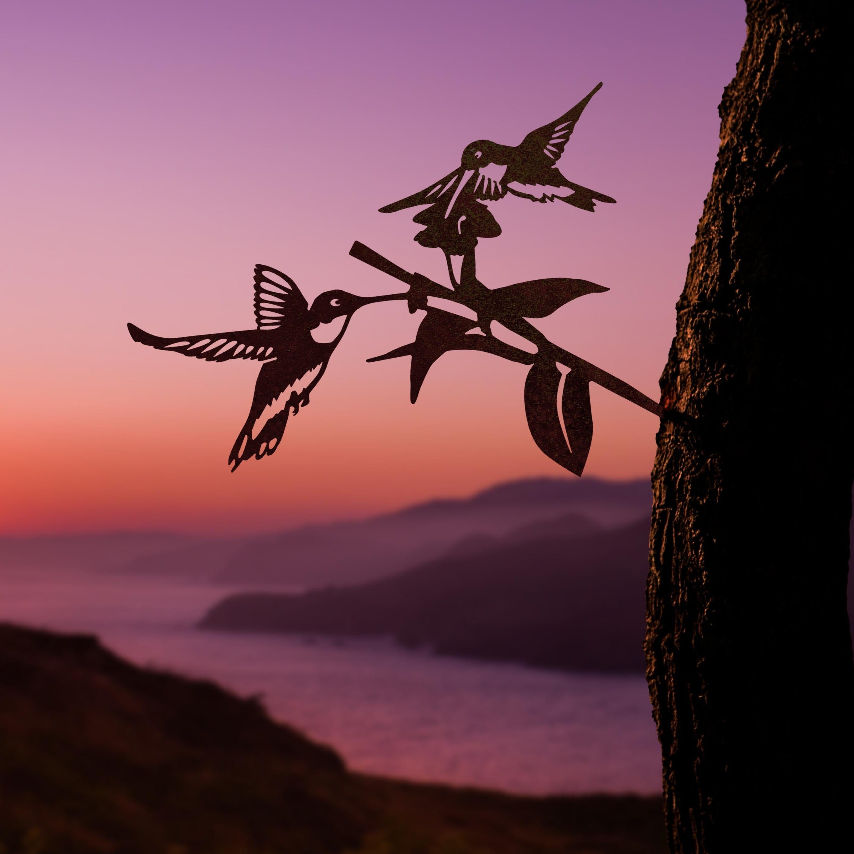 Silhouettes of a cardinal, hummingbird, and owl from the Metal Bird Garden Art collection perch on a branch against a vibrant sunset, ocean, and mountains—an ideal gift for bird lovers.