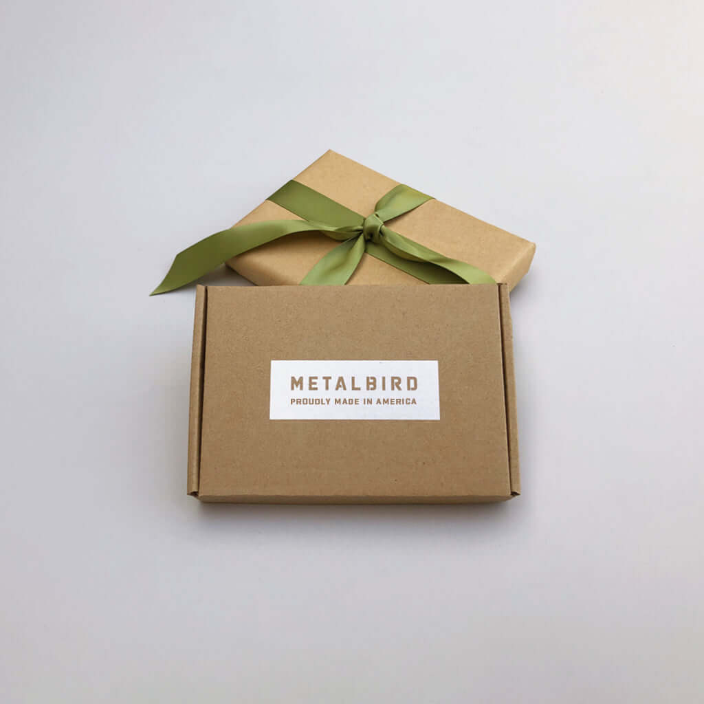Two brown gift boxes, one with a green ribbon and the other labeled METALBIRD PROUDLY MADE IN AMERICA, sit on a light gray surface—making this Baby Bird Bundle a perfect gift for unique yard art enthusiasts.