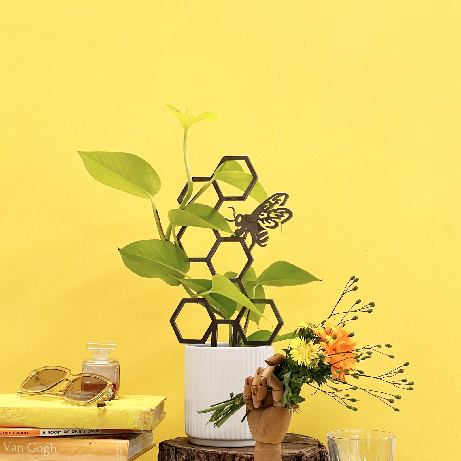 The Honeybee Plant Trellis, featuring a hexagonal honeycomb and butterfly silhouette design, supports a potted green plant on a yellow surface near books, sunglasses, and a wooden hand with flowers against a yellow wall.