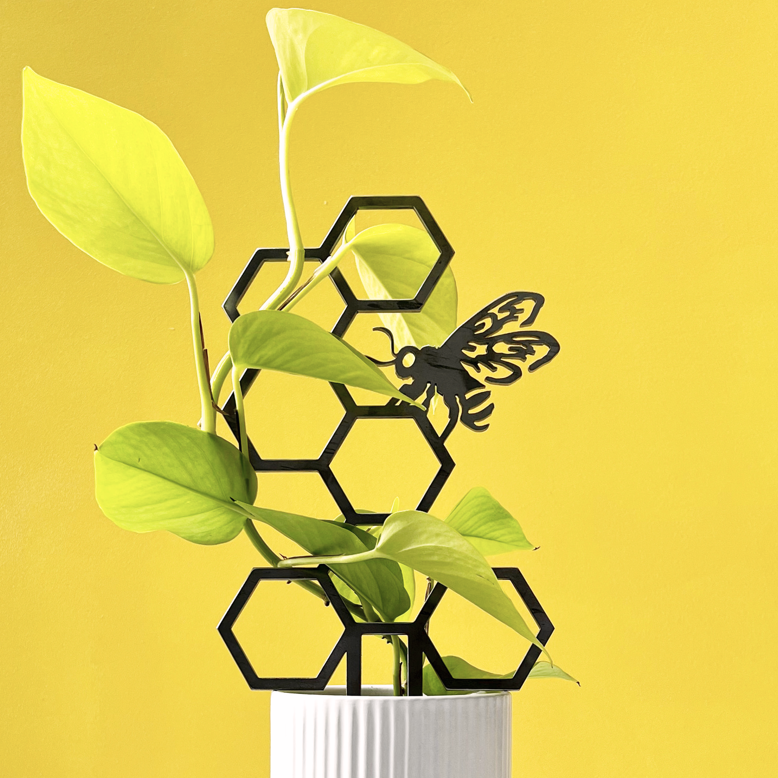 The Honeybee Plant Trellis features a green plant growing through a black corten steel honeycomb and bee sculpture in a white textured pot, set against a yellow background—an eye-catching metal gift for any space.