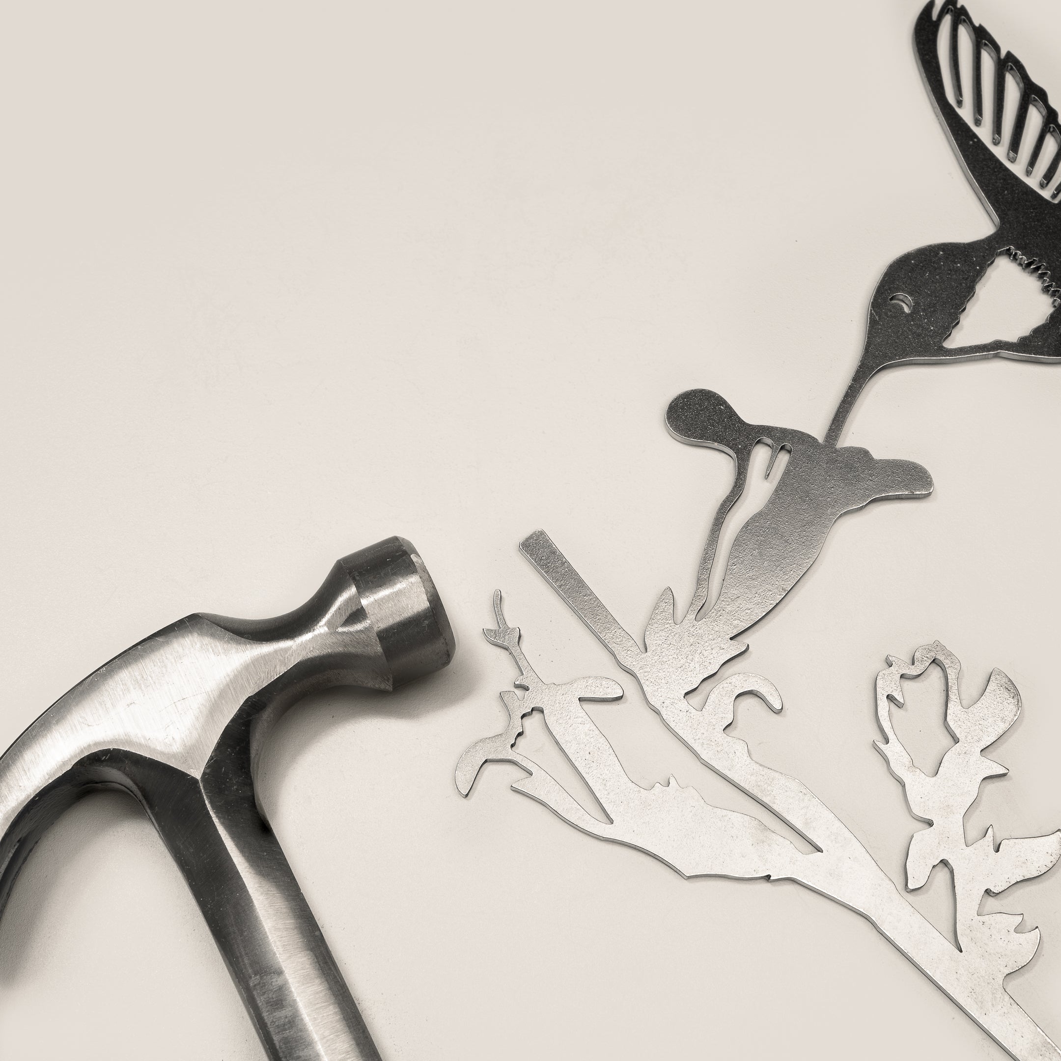 A metal hammer rests beside the Hummingbird Mom & Mini Set—intricately cut Corten steel hummingbird, flower, and branch accents—making a unique garden gift, displayed on a light surface.