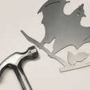The Bat features a corten steel silhouette of a bat with outstretched wings perched on a branch with two leaves, creating striking yard art. A silver hammer is placed nearby, completing the look against a light background.