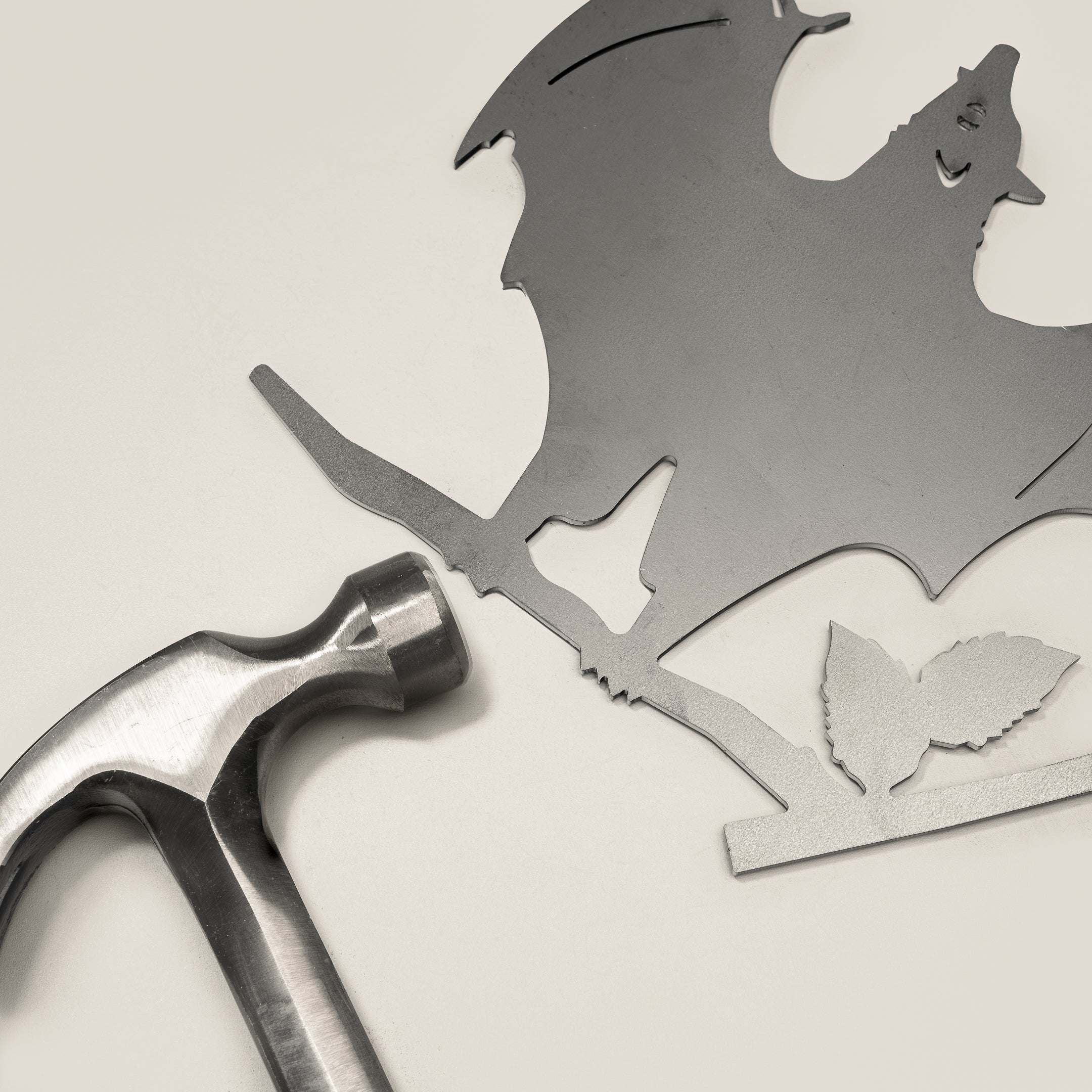 The Bat features a corten steel silhouette of a bat with outstretched wings perched on a branch with two leaves, creating striking yard art. A silver hammer is placed nearby, completing the look against a light background.