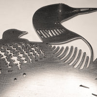 The "Common Loon" metal artwork features two loons, large and small, with cut-out feather details. Crafted from Corten steel, this piece honors Ontario's iconic bird with striking detail against a light background.
