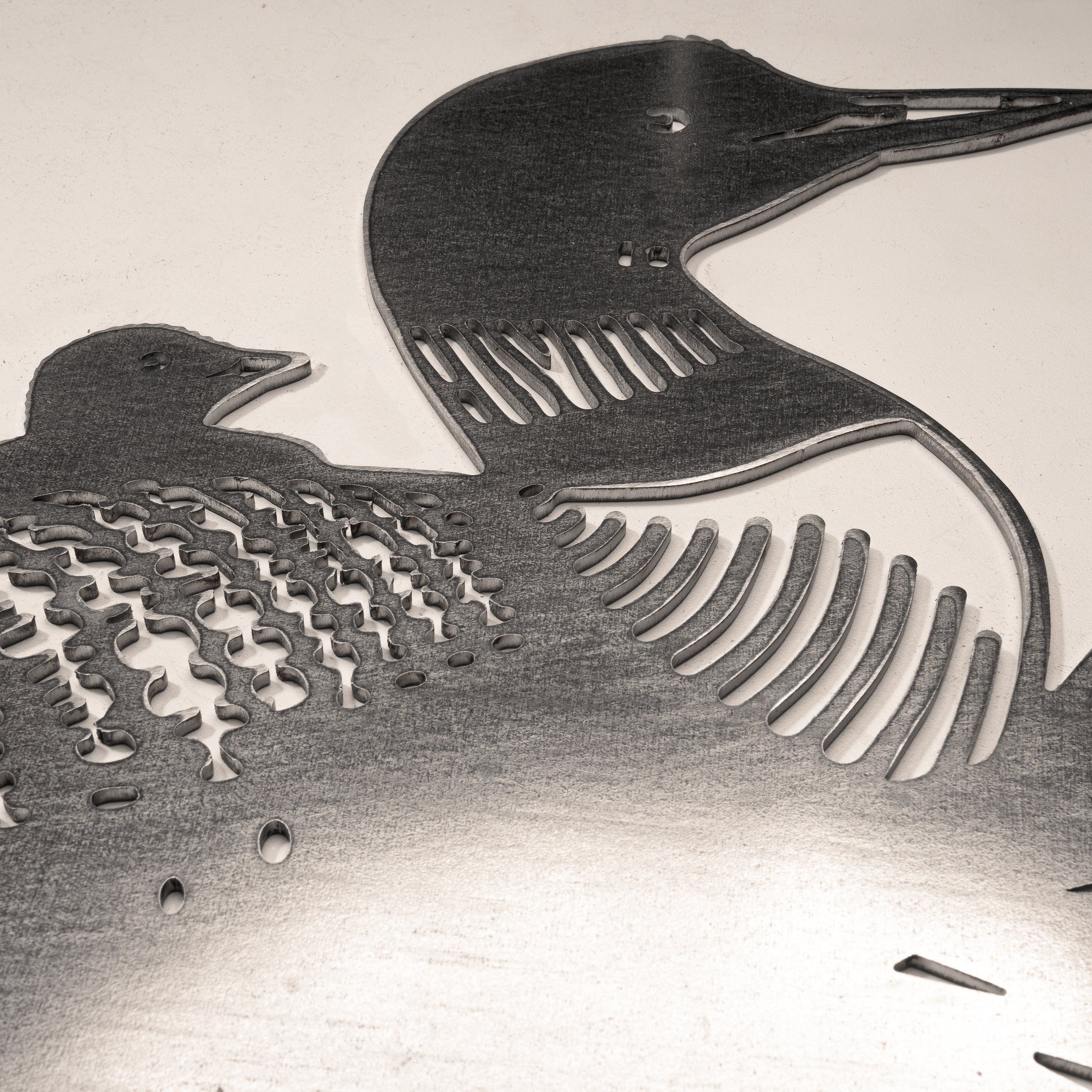The "Common Loon" metal artwork features two loons, large and small, with cut-out feather details. Crafted from Corten steel, this piece honors Ontario's iconic bird with striking detail against a light background.