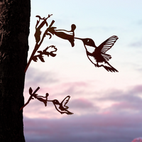 The Hummingbird Mom & Mini Set features two corten steel hummingbirds drinking from flowers on a tree trunk, with a pastel sunset sky—perfect as a hummingbird garden gift.