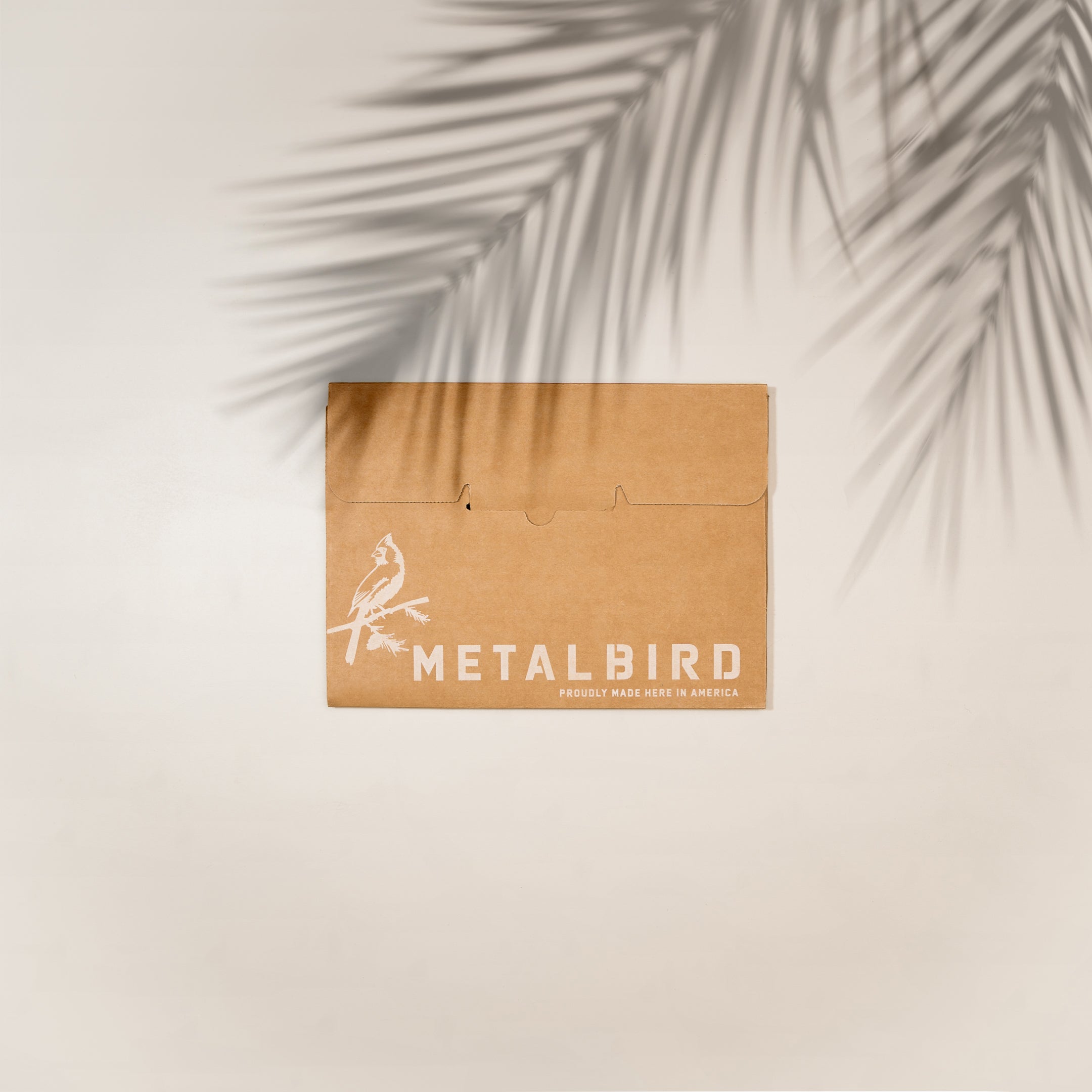 A brown box with the Metalbird and bird logo rests on a light surface, palm leaf shadows above—ideal for gifting the handcrafted Ruby-Throated Hummingbird decoration.