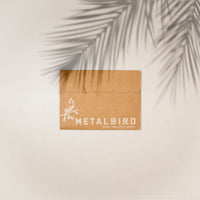 A brown METALBIRD box with a bird illustration sits on a sunlit surface cast with palm shadows—ideal for gifting the Hummingbird Mom & Mini Set, a delightful corten steel hummingbird garden gift.