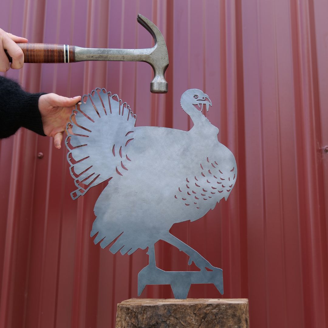 A person holds a hammer above the Turkey, a premium metal cutout shaped like a turkey on a wooden block, set against a red corrugated wall—a unique gift idea.