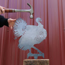 A person holds a hammer above the Turkey, a premium metal cutout shaped like a turkey on a wooden block, set against a red corrugated wall—a unique gift idea.