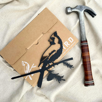 The Cardinal Black, a powder-coated cardinal silhouette perched on branches, is displayed next to a bird-illustrated brown envelope and a hammer with a brown handle on light-colored fabric.