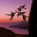 Silhouettes of a cardinal, hummingbird, and owl from the Metal Bird Garden Art collection perch on a branch against a vibrant sunset, ocean, and mountains—an ideal gift for bird lovers.