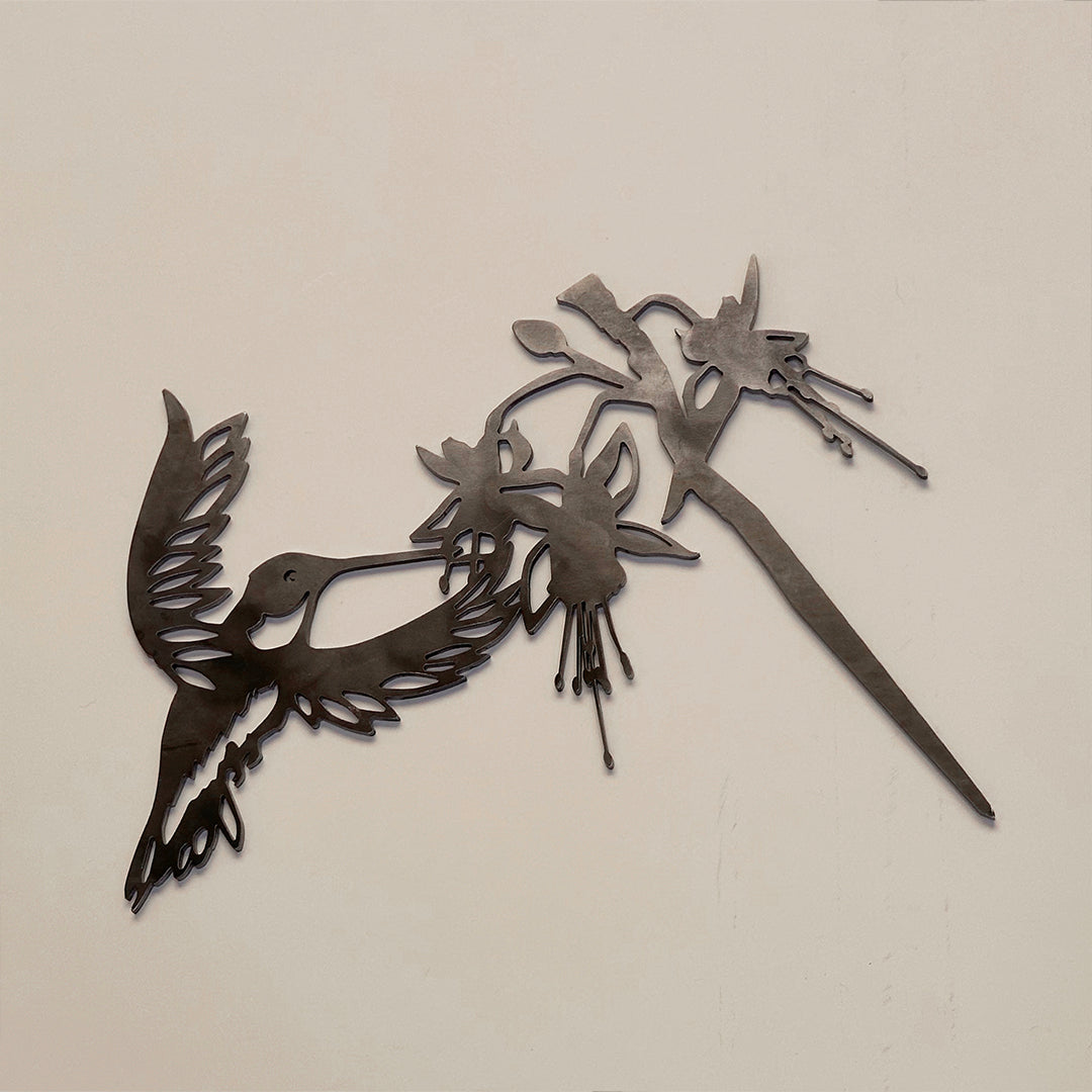 The Ruby-Throated Hummingbird metal wall art features a handcrafted silhouette of the bird with spread wings, reaching toward flowers, and is shown mounted on a plain beige wall.