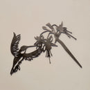 The Ruby-Throated Hummingbird metal wall art features a handcrafted silhouette of the bird with spread wings, reaching toward flowers, and is shown mounted on a plain beige wall.