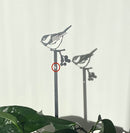 The Metalbird Seconds - Chickadee Plant Stake casts a sharp shadow on a white wall. Sunlight highlights the bird perched above berries, with green leaves and a red circle adding detail below.