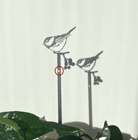 The Metalbird Seconds - Chickadee Plant Stake casts a sharp shadow on a white wall. Sunlight highlights the bird perched above berries, with green leaves and a red circle adding detail below.