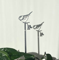 The Metalbird Seconds - Chickadee Plant Stake stands among green leaves, casting a clear bird silhouette perched on a branch against a plain white wall.