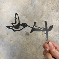 A hand holds a Corten steel Hummingbird Mom & Mini Set cutout, shaped like birds drinking from flowers against a concrete floor—the perfect garden gift for hummingbird lovers.