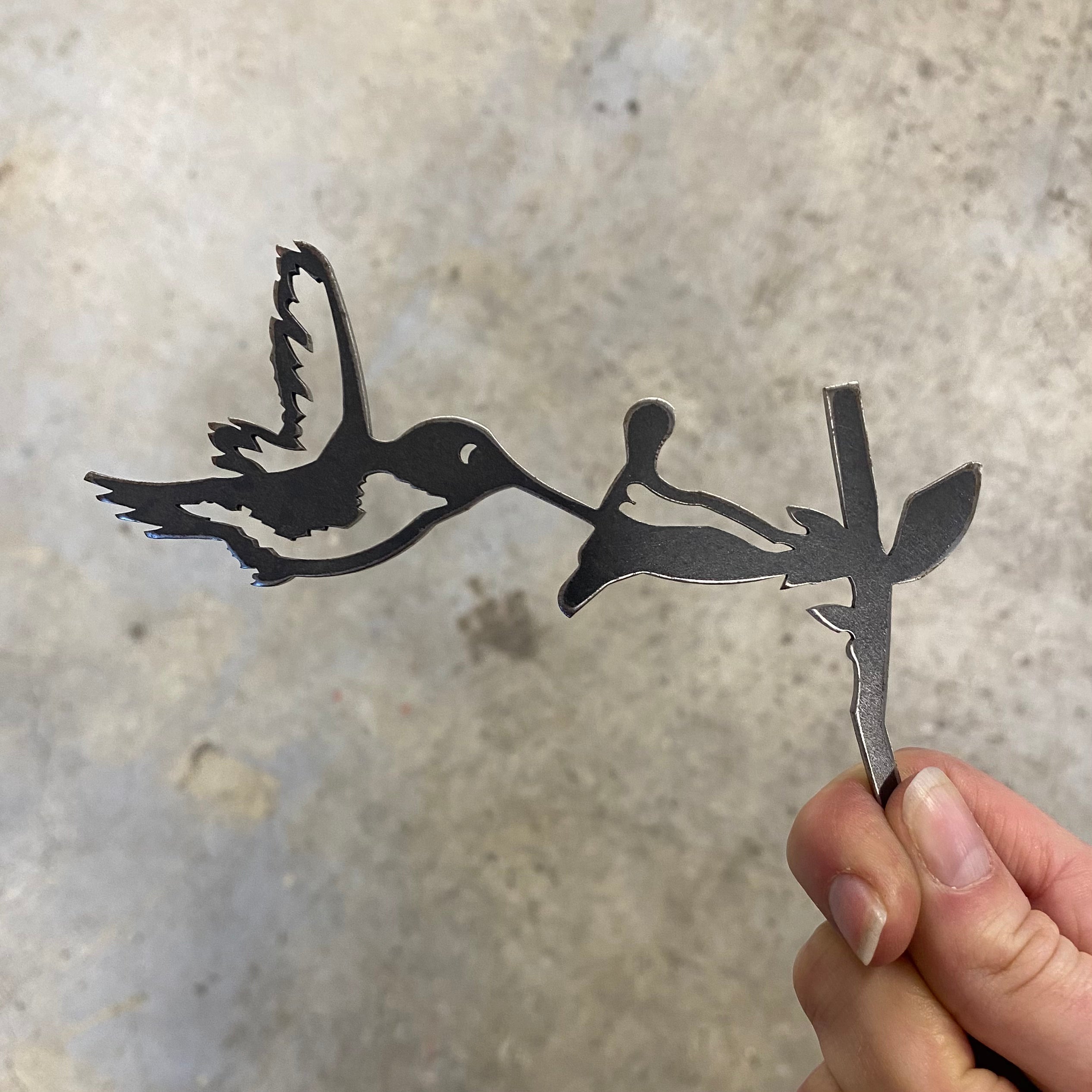 A hand holds the Mini Metal Bird Garden Art – Hummingbird, a steel cutout with detailed wings and beak, adding unique decor for bird lovers against a light gray concrete background. Also available as Cardinal and Owl.