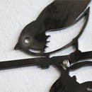 A close-up of the Metalbird Seconds - Chickadee, a corten steel wall art piece with a stylized chickadee silhouette perched on a branch, featuring delicate cut-out details and mounted on a textured white wall.