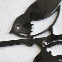 A close-up of the Metalbird Seconds - Hummingbird, a premium black metal wall art featuring a hummingbird perched on a branch, mounted on a white textured wall—an ideal gift for any decor enthusiast.