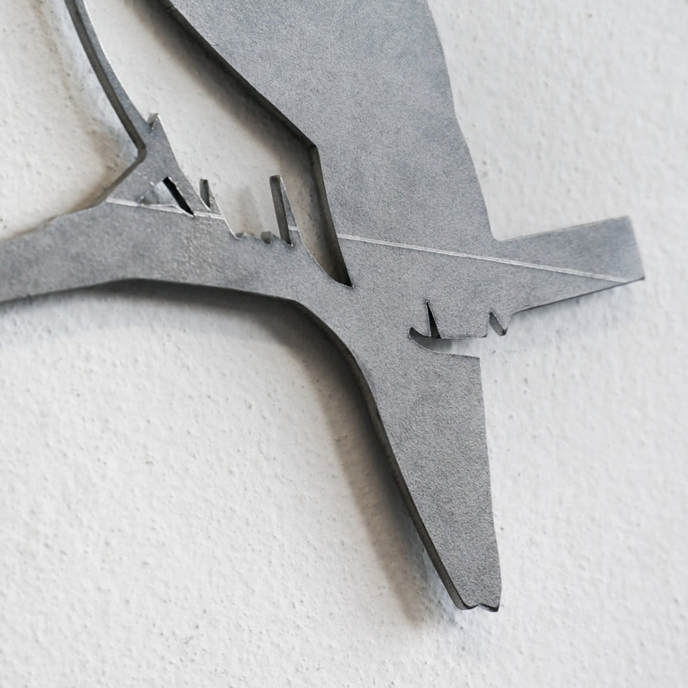 A close-up of the Metalbird Seconds - Chickadee, a minimalist metal wall art piece featuring a bird silhouette perched on a branch, displayed on a light-colored textured wall.