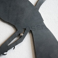 A close-up of the Metalbird Seconds - Chickadee metal wall art, shaped like a bird silhouette and mounted on a white wall. Textured surface, visible screw, and partially engraved letters are shown.