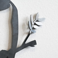 A close-up of the Metalbird Seconds - Cardinal, a corten steel wall decoration featuring part of a curved shape holding an olive branch, mounted on a light textured wall.