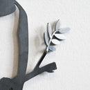 A close-up of the Metalbird Seconds - Canada Jay wall decoration reveals part of a branch with leaves on a textured white wall. The matte gray finish features subtle imperfections, giving the design unique character.