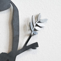 A close-up of the Metalbird Seconds - Canada Jay wall decoration reveals part of a branch with leaves on a textured white wall. The matte gray finish features subtle imperfections, giving the design unique character.