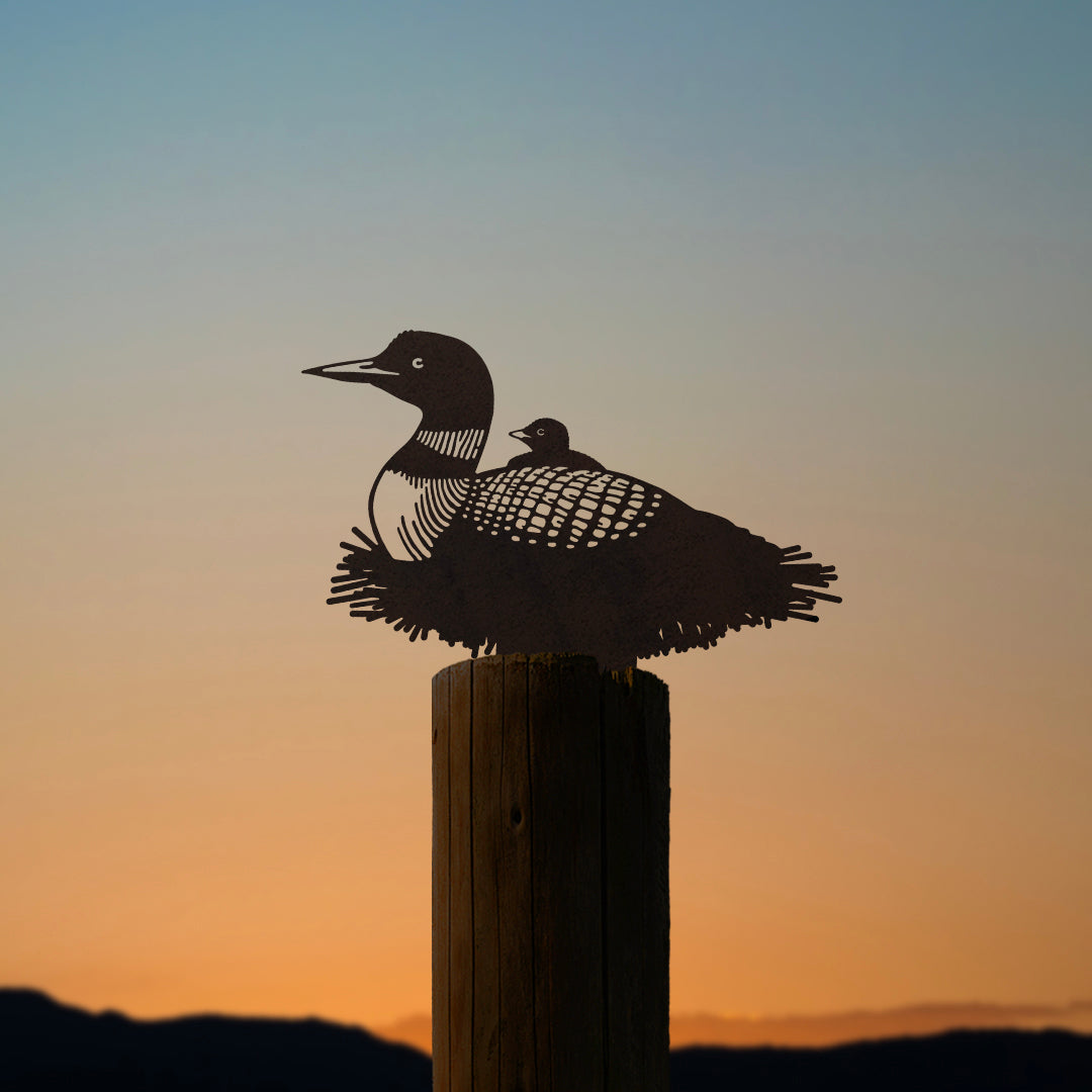The Common Loon is a metal silhouette of this iconic Ontario bird with a chick, crafted from Corten steel and mounted on wood. Set against an orange and blue sunset, it captures the beauty of the species in a unique decorative piece.