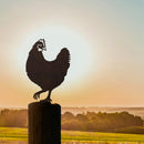 The Rhode Island Red, a Corten steel hen inspired by the state bird, is mounted on a wooden post with scenic fields and trees in the background at sunset.