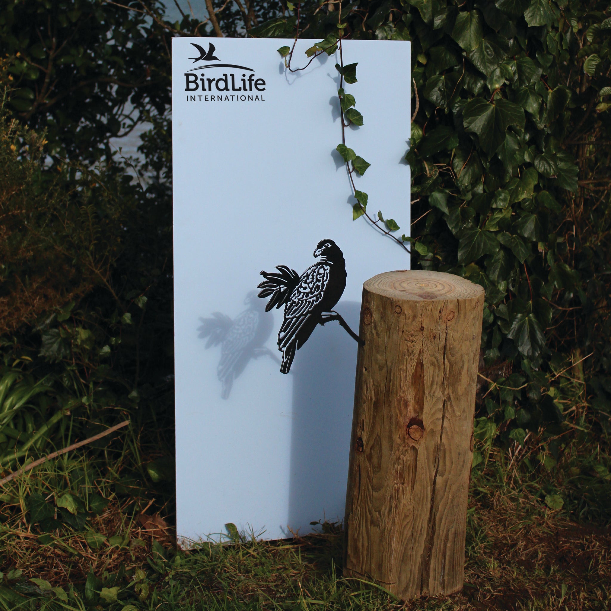 A Manumea sign featuring the BirdLife International logo and a black bird illustration stands outdoors amid greenery beside a wooden log pedestal, with a vine draped over it.