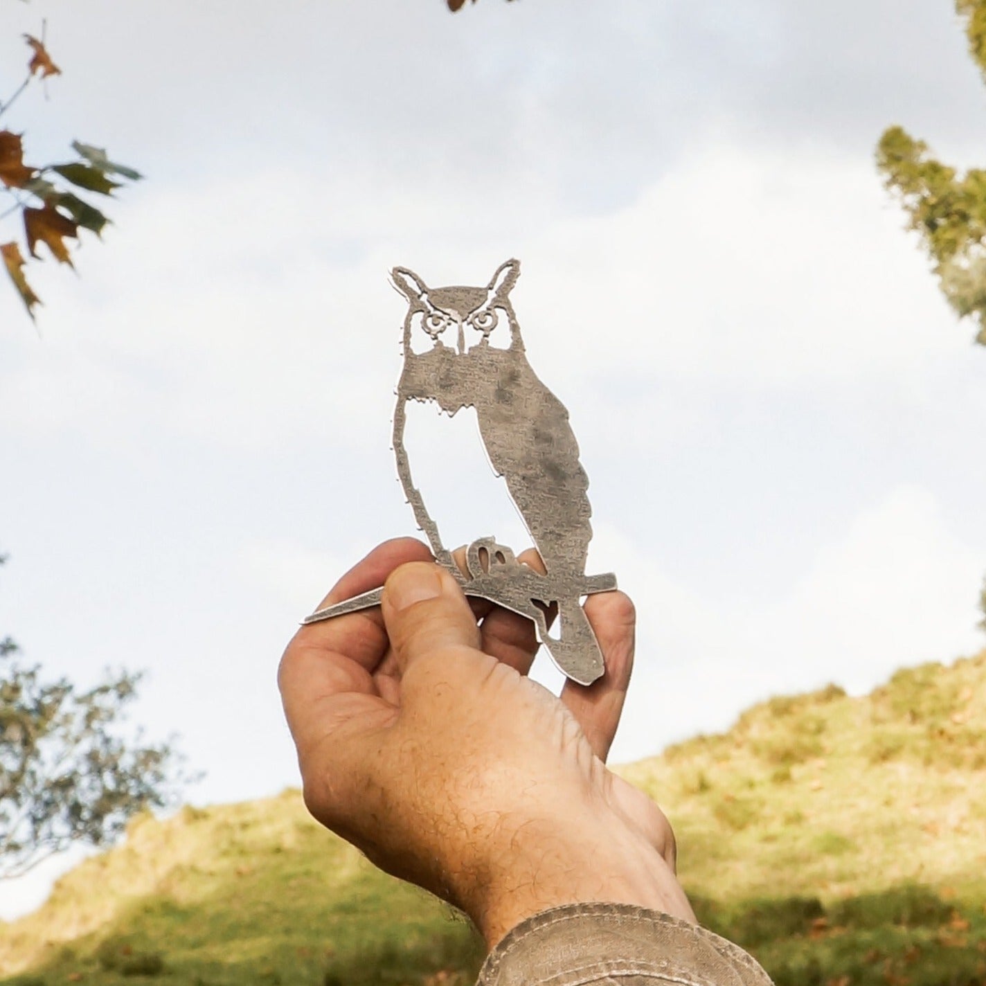 A hand displays the Mini Metal Bird Garden Art – Owl, an ideal gift for bird lovers and perfect garden decor, with a grassy hill and cloudy sky in the background.