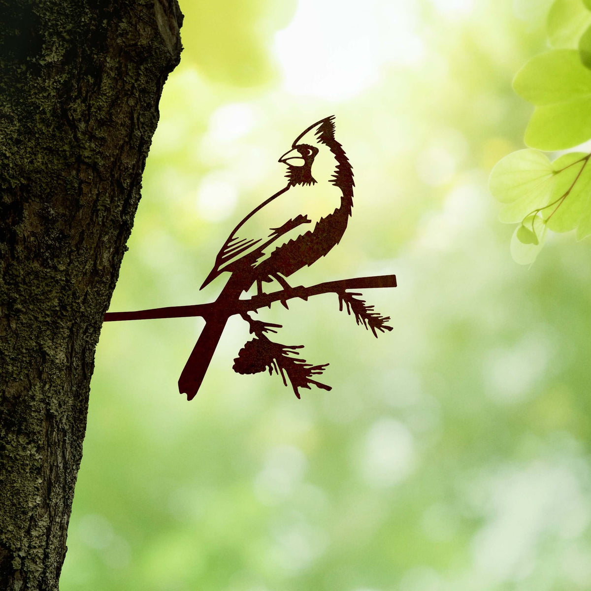 Cardinal steel silhouette mounted beside a tree trunk against green leaves.