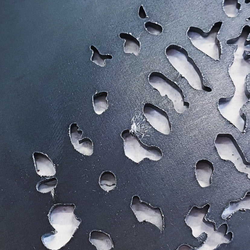 Close-up of the Metalbird Seconds - Pileated Woodpecker: dark corten steel with irregular, organic-shaped cutouts revealing a lighter background and forming an abstract, textured pattern of varying silhouettes.
