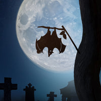 The Bat, crafted in corten steel, hangs upside down from a tree branch with a full moon and gravestones behind—perfect yard art for a spooky scene or as a unique gift.