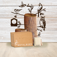 A wooden stand holds black metal bird silhouettes—an owl and songbirds. Nearby, brown envelopes labeled METALBIRD, the Christmas Favorites Gift Pack, and a small branded box rest on a light wood surface.