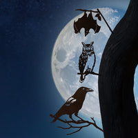 Corten steel crow, owl, and bat silhouettes perch on a tree branch under a full moon. Spooky garden art gift.