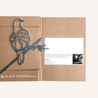 A brown cardboard package contains a Turtle Dove Corten steel bird sculpture sitting on a branch to the left. To the right, there's a white card with printed text and a black-and-white photo. Recycling symbols are at the bottom left.