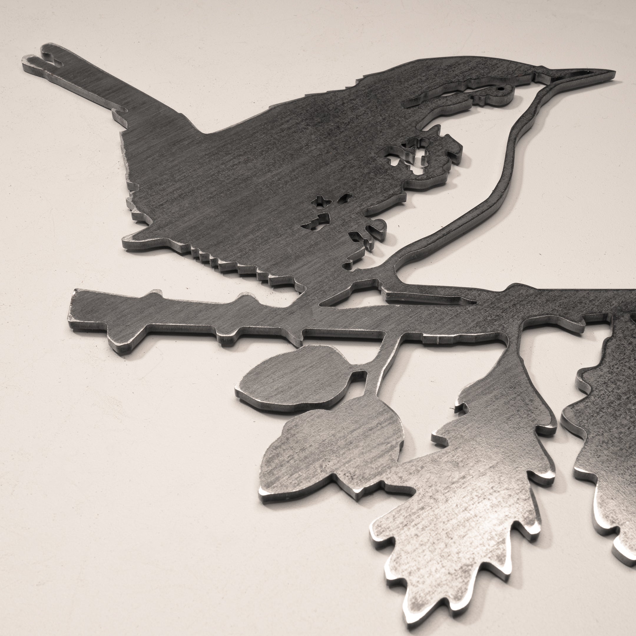 A corten steel silhouette cutout named "Wren" features a bird perched on a leafy branch, displayed on a light surface with a soft, sepia tone—ideal as a distinctive gift or delightful decor accent.