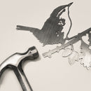 A metal hammer rests beside the Wren, an intricately cut corten steel silhouette of a bird on a branch, both set against a plain light background.