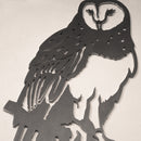 The Barn Owl is a black metal wall art piece made from durable Corten steel, with cut-out feather and face details, shown perched on a branch against a light textured wall—perfect as a hunting gift.
