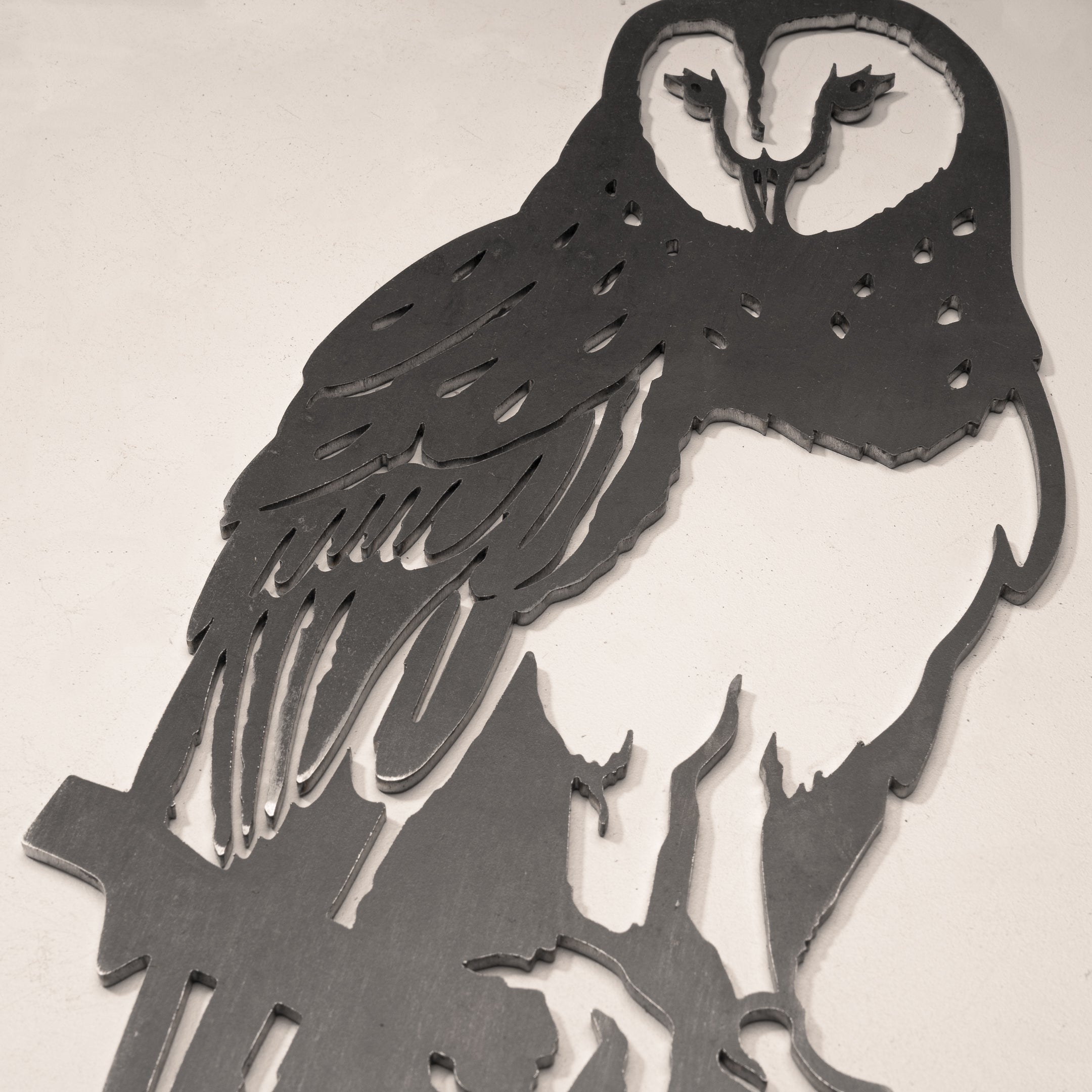 The Barn Owl is a black metal wall art piece made from durable Corten steel, with cut-out feather and face details, shown perched on a branch against a light textured wall—perfect as a hunting gift.