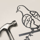 A metal hammer sits beside the Turtle Dove, a slim and elegant Corten steel bird ornament showing a dove on a branch, both set on a light surface.
